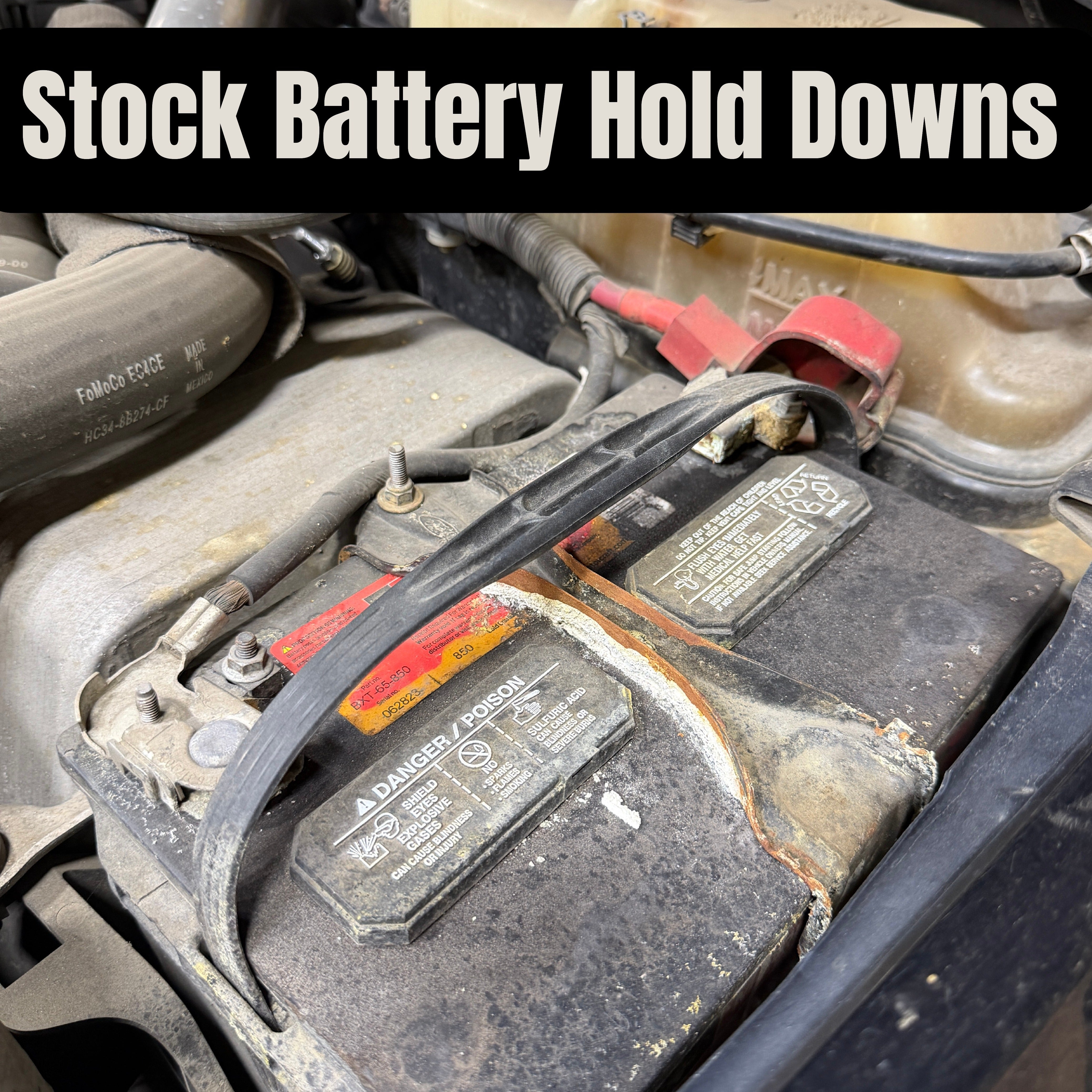 Close-up of a stock vehicle battery with corroded terminals and a plastic hold-down strap, labeled "Stock Battery Hold Downs"
